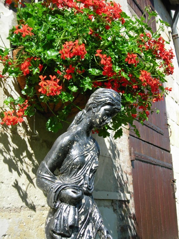 Stone lady statue beside red geraniums at Moulin St Blaise, Chahaignes, France